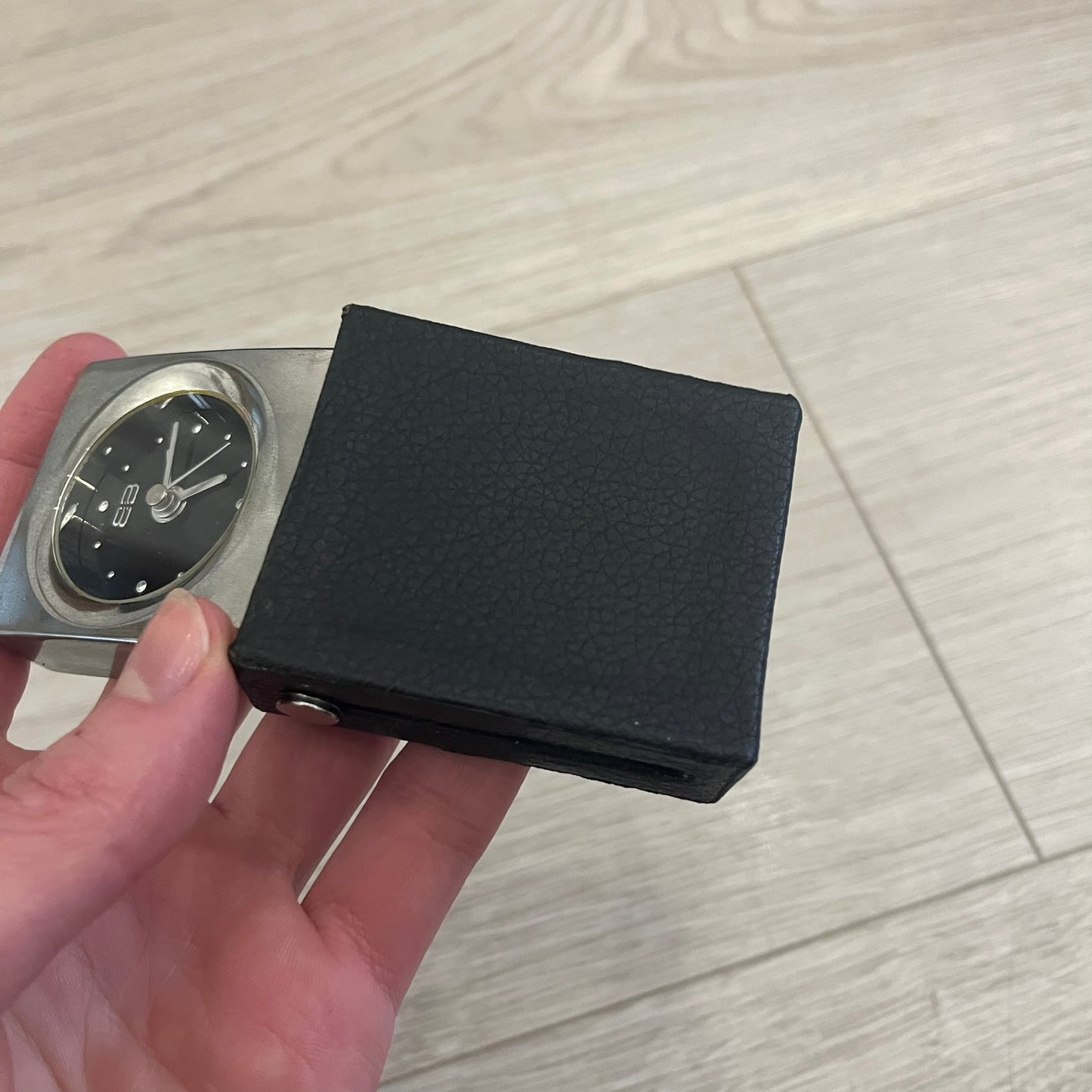 Compact silver-tone metal travel clock with engraved Balenciaga logo. Features a black dial with BB logo and silver-colored hands, plus “Siofor 1000” engraving on the top. Battery-powered and housed in a removable faux leather cover secured by movable side fasteners. Likely produced as a promotional or corporate gift piece.