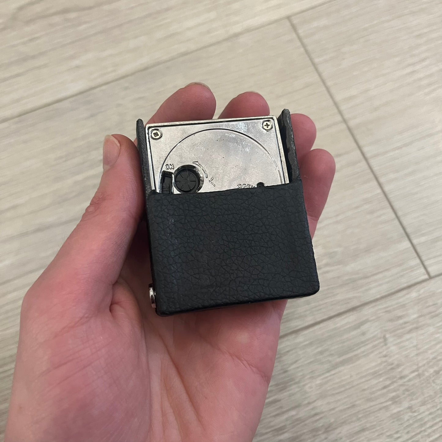 Compact silver-tone metal travel clock with engraved Balenciaga logo. Features a black dial with BB logo and silver-colored hands, plus “Siofor 1000” engraving on the top. Battery-powered and housed in a removable faux leather cover secured by movable side fasteners. Likely produced as a promotional or corporate gift piece.