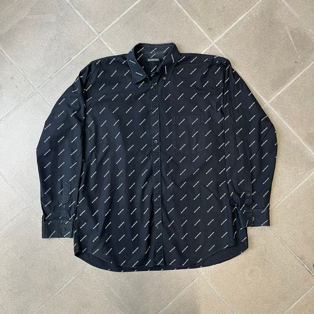 Balenciaga SS18 Black All-Over Logo Shirt is a black button-up made in Italy, featuring a diagonal all-over pattern of small white Balenciaga logo text and black buttons branded with Balenciaga. It has the archetype tag from the Spring/Summer 2018 collection.