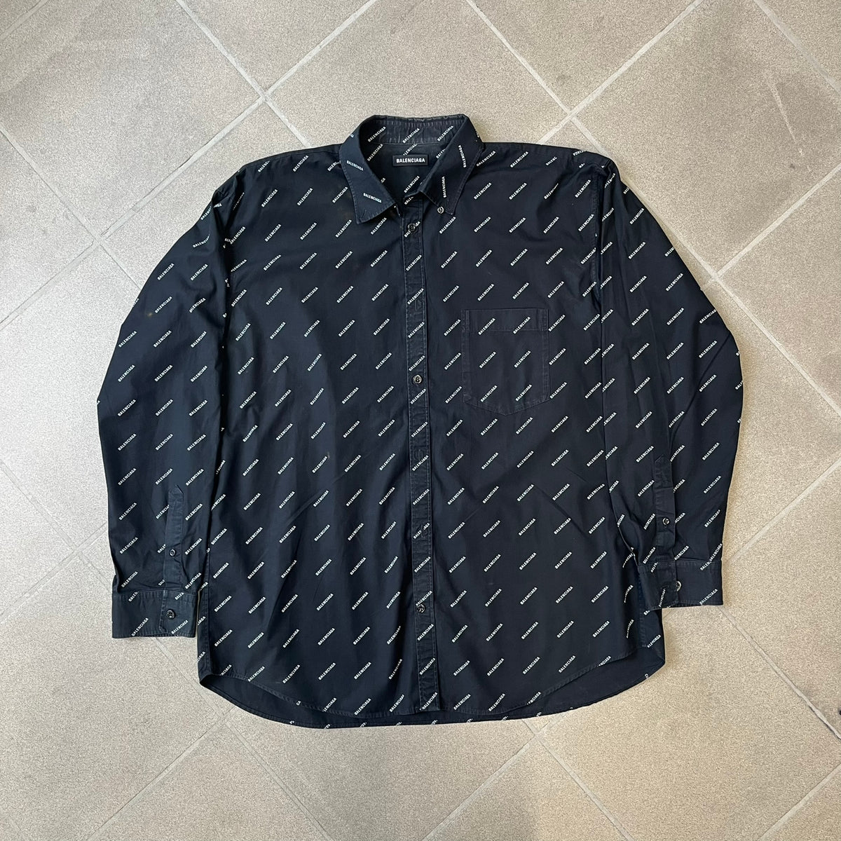 Balenciaga SS18 Black All-Over Logo Shirt is a black button-up made in Italy, featuring a diagonal all-over pattern of small white Balenciaga logo text and black buttons branded with Balenciaga. It has the archetype tag from the Spring/Summer 2018 collection.