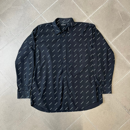 Balenciaga SS18 Black All-Over Logo Shirt is a black button-up made in Italy, featuring a diagonal all-over pattern of small white Balenciaga logo text and black buttons branded with Balenciaga. It has the archetype tag from the Spring/Summer 2018 collection.