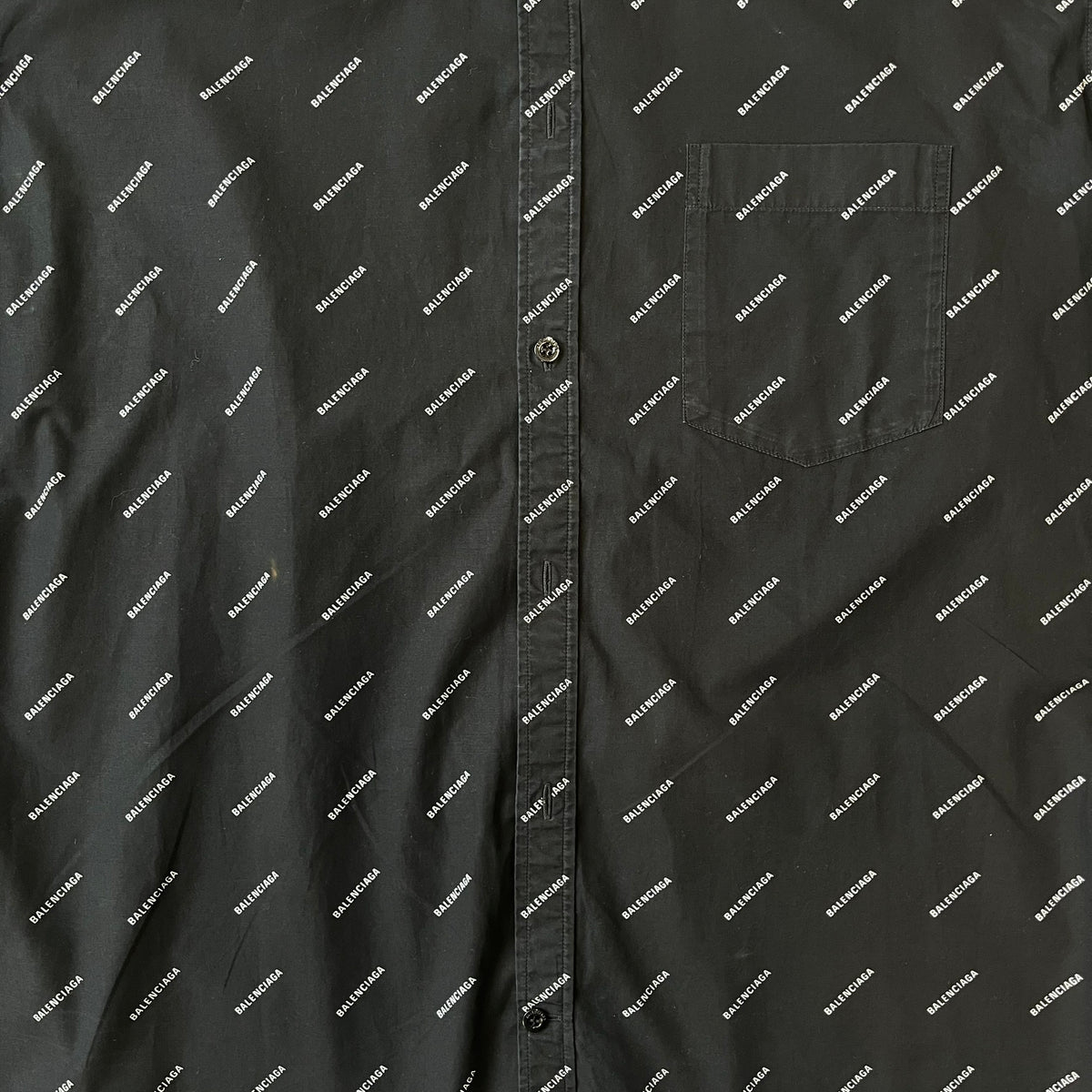 Balenciaga SS18 Black All-Over Logo Shirt is a black button-up made in Italy, featuring a diagonal all-over pattern of small white Balenciaga logo text and black buttons branded with Balenciaga. It has the archetype tag from the Spring/Summer 2018 collection.