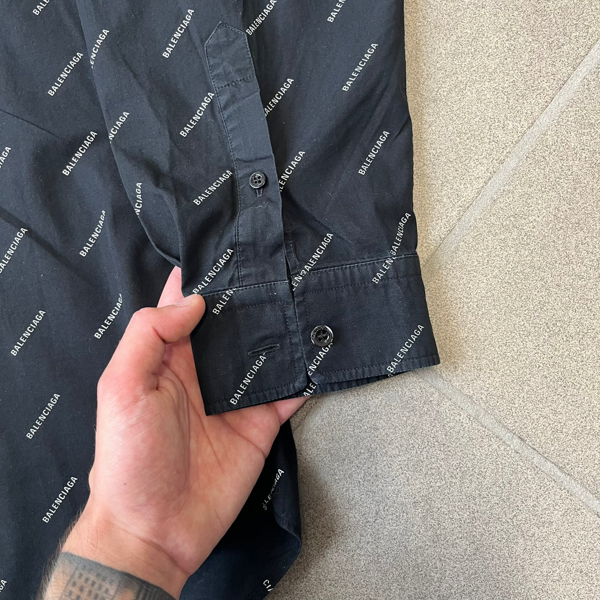 Balenciaga SS18 Black All-Over Logo Shirt is a black button-up made in Italy, featuring a diagonal all-over pattern of small white Balenciaga logo text and black buttons branded with Balenciaga. It has the archetype tag from the Spring/Summer 2018 collection.