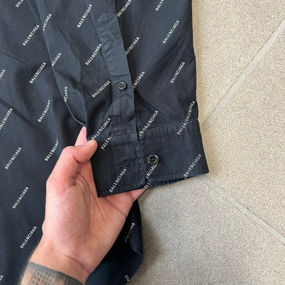 Balenciaga SS18 Black All-Over Logo Shirt is a black button-up made in Italy, featuring a diagonal all-over pattern of small white Balenciaga logo text and black buttons branded with Balenciaga. It has the archetype tag from the Spring/Summer 2018 collection.