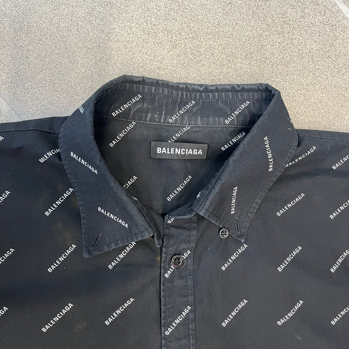 Balenciaga SS18 Black All-Over Logo Shirt is a black button-up made in Italy, featuring a diagonal all-over pattern of small white Balenciaga logo text and black buttons branded with Balenciaga. It has the archetype tag from the Spring/Summer 2018 collection.