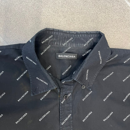 Balenciaga SS18 Black All-Over Logo Shirt is a black button-up made in Italy, featuring a diagonal all-over pattern of small white Balenciaga logo text and black buttons branded with Balenciaga. It has the archetype tag from the Spring/Summer 2018 collection.