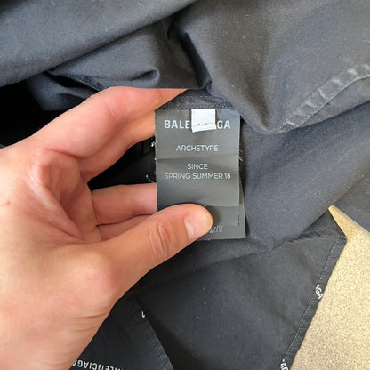 Balenciaga SS18 Black All-Over Logo Shirt is a black button-up made in Italy, featuring a diagonal all-over pattern of small white Balenciaga logo text and black buttons branded with Balenciaga. It has the archetype tag from the Spring/Summer 2018 collection.
