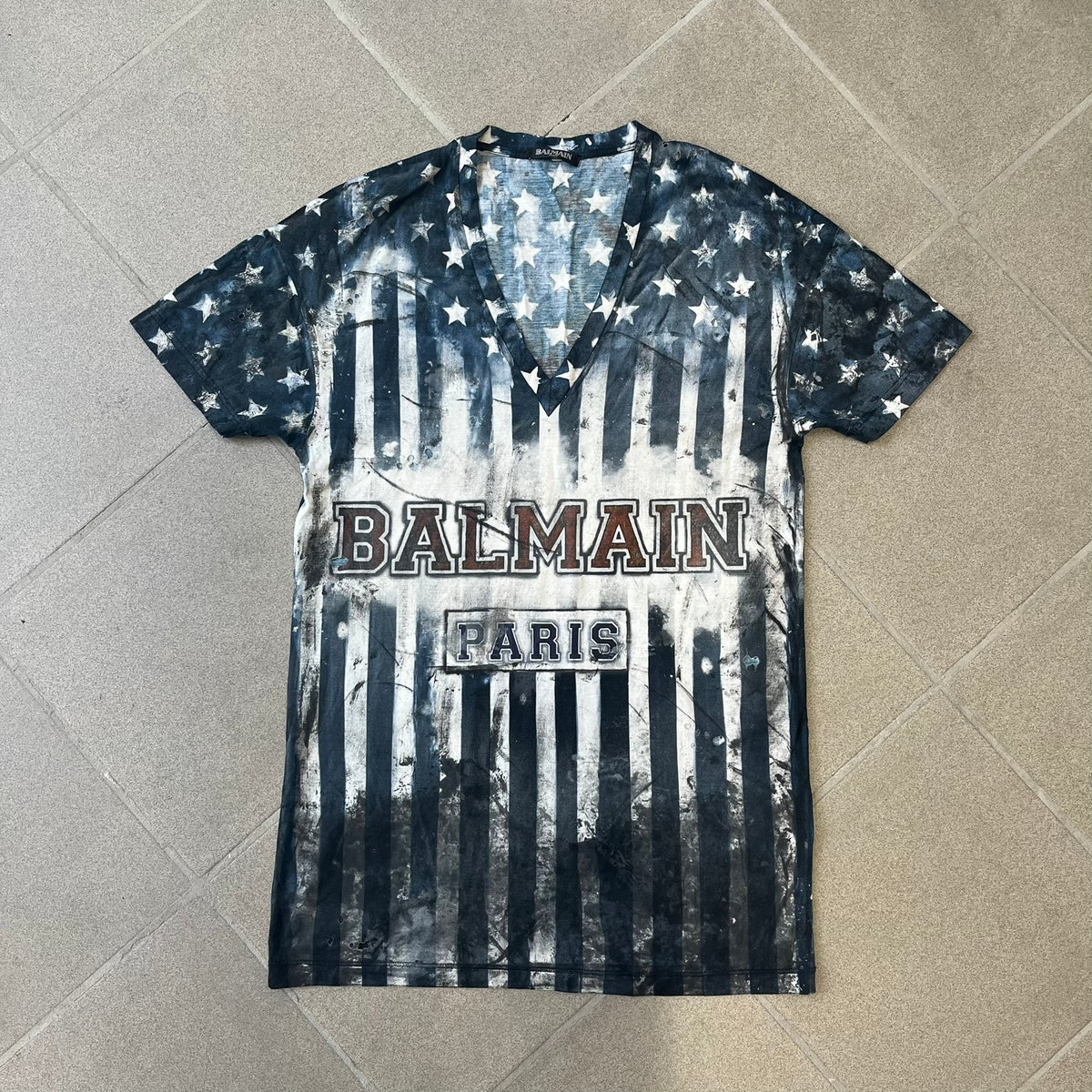 Balmain 2018 American Flag V-Neck Tee is a 100% cotton tee featuring an all-over blue and white American flag print, with stars at the top and stripes through the middle and bottom, and a red Balmain logo centered on the design. It has an intentionally distressed finish with a dirtied color treatment and holes throughout.