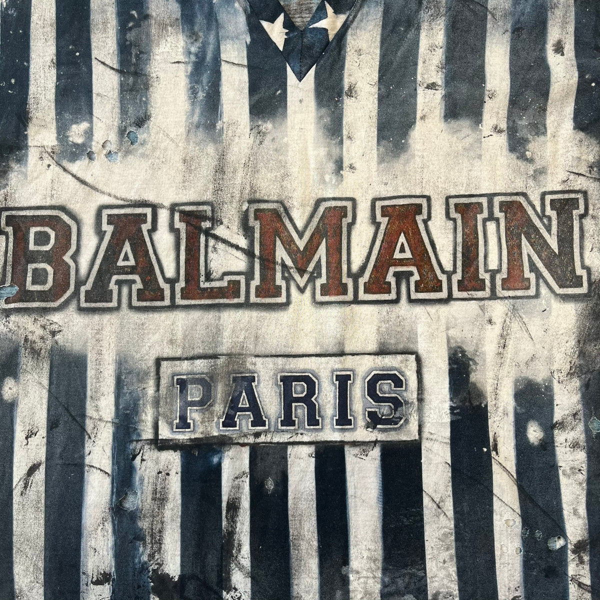 Balmain 2018 American Flag V-Neck Tee is a 100% cotton tee featuring an all-over blue and white American flag print, with stars at the top and stripes through the middle and bottom, and a red Balmain logo centered on the design. It has an intentionally distressed finish with a dirtied color treatment and holes throughout.