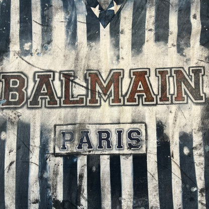 Balmain 2018 American Flag V-Neck Tee is a 100% cotton tee featuring an all-over blue and white American flag print, with stars at the top and stripes through the middle and bottom, and a red Balmain logo centered on the design. It has an intentionally distressed finish with a dirtied color treatment and holes throughout.