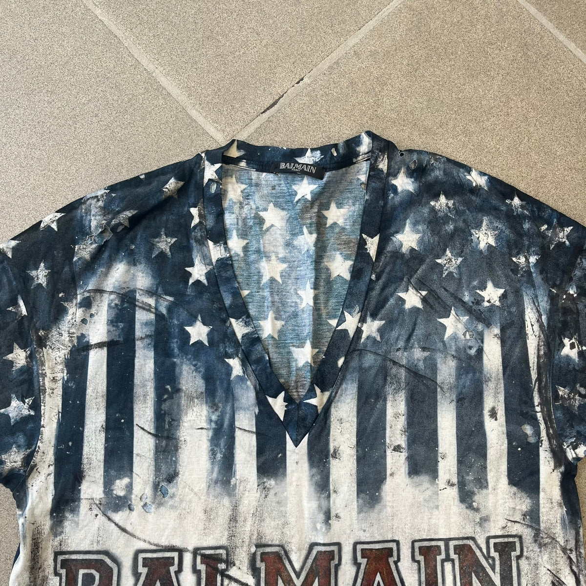 Balmain 2018 American Flag V-Neck Tee is a 100% cotton tee featuring an all-over blue and white American flag print, with stars at the top and stripes through the middle and bottom, and a red Balmain logo centered on the design. It has an intentionally distressed finish with a dirtied color treatment and holes throughout.