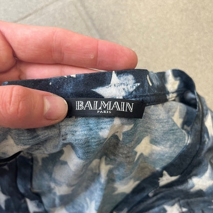 Balmain 2018 American Flag V-Neck Tee is a 100% cotton tee featuring an all-over blue and white American flag print, with stars at the top and stripes through the middle and bottom, and a red Balmain logo centered on the design. It has an intentionally distressed finish with a dirtied color treatment and holes throughout.