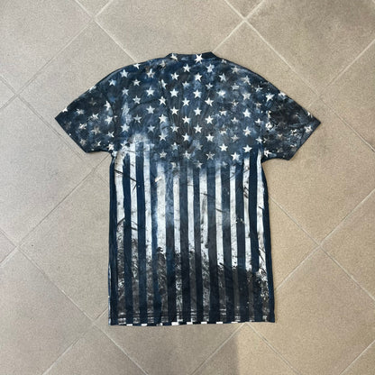 Balmain 2018 American Flag V-Neck Tee is a 100% cotton tee featuring an all-over blue and white American flag print, with stars at the top and stripes through the middle and bottom, and a red Balmain logo centered on the design. It has an intentionally distressed finish with a dirtied color treatment and holes throughout.