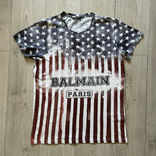 Balmain SS18 t-shirt in 100% cotton featuring a bold full-print USA flag on both sides. The front is accented with “Balmain Paris” text at the stomach, combining patriotic imagery with the brand’s signature luxury touch. A rare and powerful piece from the 2018 runway season.