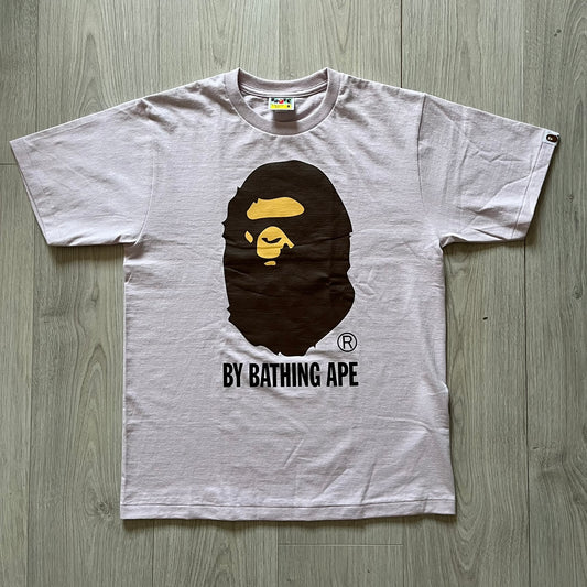 Bape tee in a pinkish/light purple shade, featuring the brand’s classic Ape Head graphic on the front. “By Bathing Ape” text sits underneath for a clean, iconic finish.