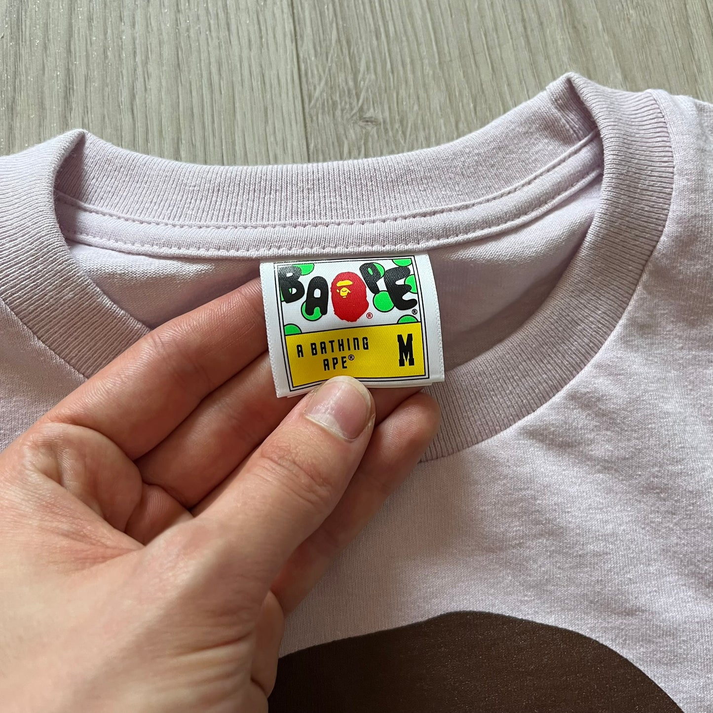 Bape tee in a pinkish/light purple shade, featuring the brand’s classic Ape Head graphic on the front. “By Bathing Ape” text sits underneath for a clean, iconic finish.