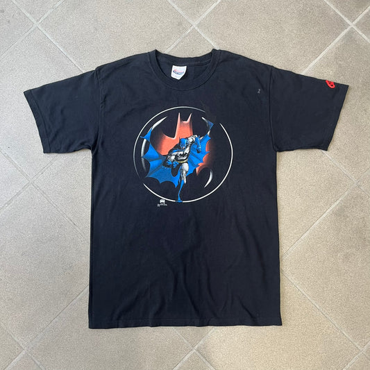 Batman 2005 Graphitti Red Logo Tee is a black T-shirt featuring a graphic of Batman in a blue suit running forward, set against a circular red Bat logo in the background. A red Graphitti logo is placed on one sleeve.