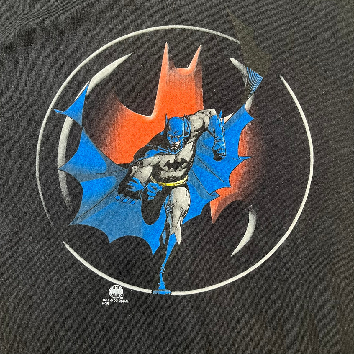 Batman 2005 Graphitti Red Logo Tee is a black T-shirt featuring a graphic of Batman in a blue suit running forward, set against a circular red Bat logo in the background. A red Graphitti logo is placed on one sleeve.