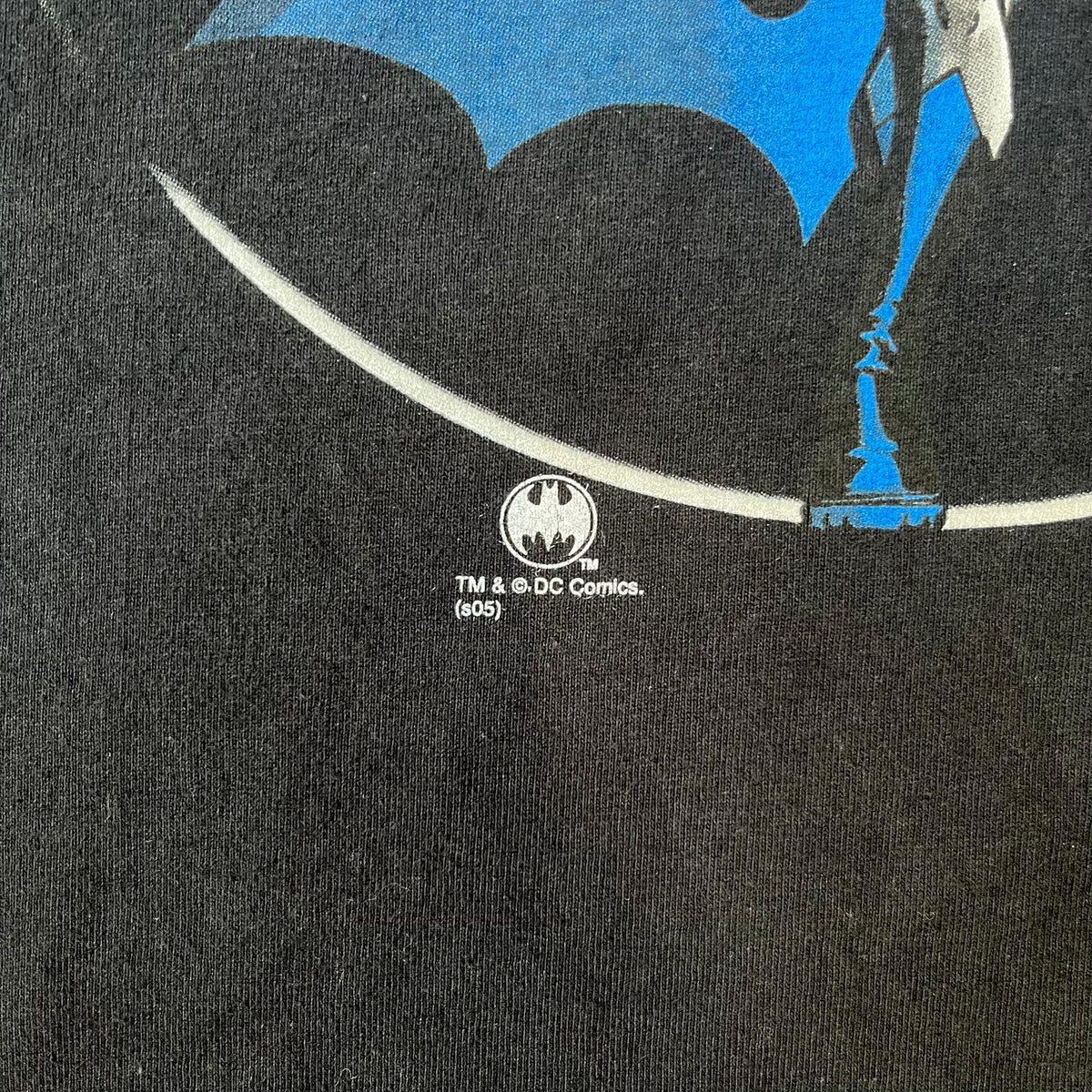 Batman 2005 Graphitti Red Logo Tee is a black T-shirt featuring a graphic of Batman in a blue suit running forward, set against a circular red Bat logo in the background. A red Graphitti logo is placed on one sleeve.