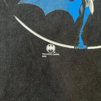 Batman 2005 Graphitti Red Logo Tee is a black T-shirt featuring a graphic of Batman in a blue suit running forward, set against a circular red Bat logo in the background. A red Graphitti logo is placed on one sleeve.