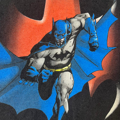 Batman 2005 Graphitti Red Logo Tee is a black T-shirt featuring a graphic of Batman in a blue suit running forward, set against a circular red Bat logo in the background. A red Graphitti logo is placed on one sleeve.