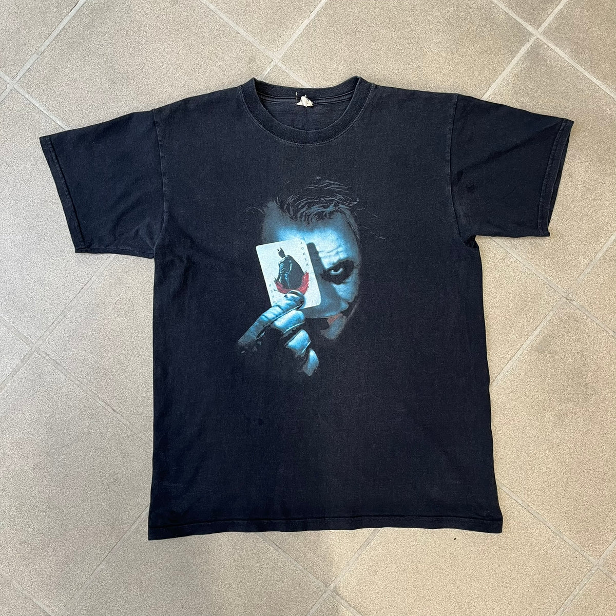 Batman The Dark Knight 2008 Joker Promo Tee features a black construction made on an Anvil blank, with Heath Ledger as the Joker holding a Batman playing card printed on the front. The upper back has The Dark Knight logo with small DC Comics 2008 licensing text underneath.
