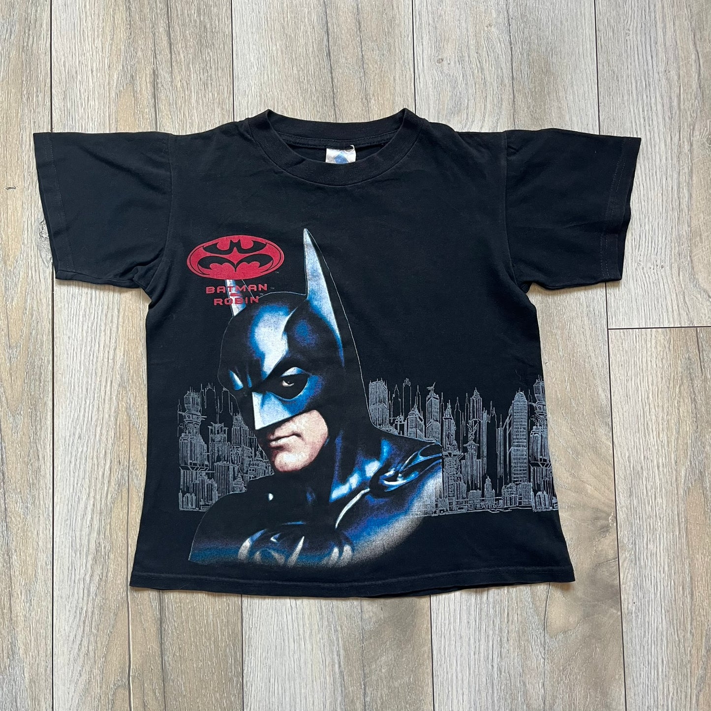 Batman &amp; Robin 1997 Mr. Freeze Promo Tee is a black T-shirt featuring a front graphic of Batman’s masked bust set against a grey city background, with a red Batman &amp; Robin logo above. The cityscape continues from the front around to the back, where a realistic Mr. Freeze graphic appears with his blue text incorporating the Batman symbol. Small 1997 licensing text is printed beneath the back graphic.