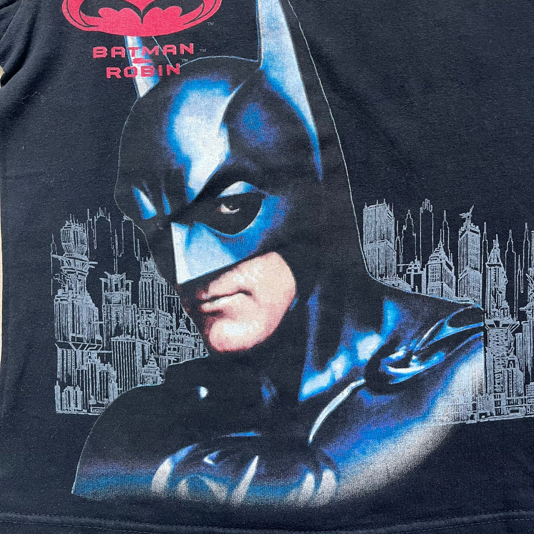 Batman &amp; Robin 1997 Mr. Freeze Promo Tee is a black T-shirt featuring a front graphic of Batman’s masked bust set against a grey city background, with a red Batman &amp; Robin logo above. The cityscape continues from the front around to the back, where a realistic Mr. Freeze graphic appears with his blue text incorporating the Batman symbol. Small 1997 licensing text is printed beneath the back graphic.
