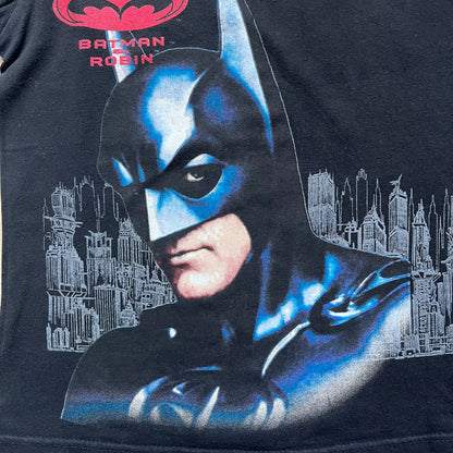 Batman &amp; Robin 1997 Mr. Freeze Promo Tee is a black T-shirt featuring a front graphic of Batman’s masked bust set against a grey city background, with a red Batman &amp; Robin logo above. The cityscape continues from the front around to the back, where a realistic Mr. Freeze graphic appears with his blue text incorporating the Batman symbol. Small 1997 licensing text is printed beneath the back graphic.