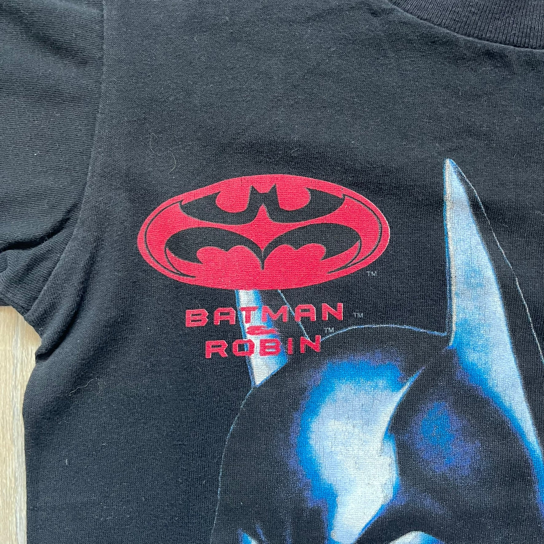 Batman &amp; Robin 1997 Mr. Freeze Promo Tee is a black T-shirt featuring a front graphic of Batman’s masked bust set against a grey city background, with a red Batman &amp; Robin logo above. The cityscape continues from the front around to the back, where a realistic Mr. Freeze graphic appears with his blue text incorporating the Batman symbol. Small 1997 licensing text is printed beneath the back graphic.