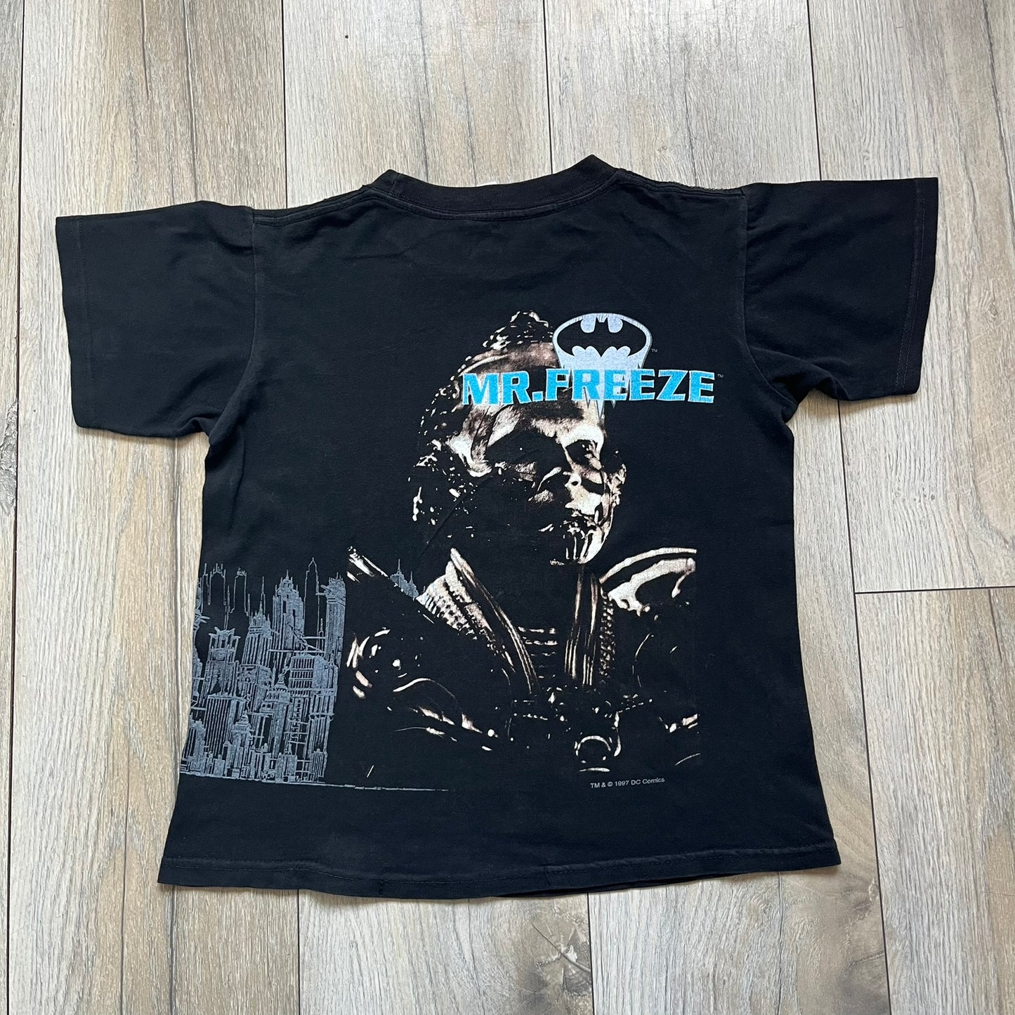 Batman &amp; Robin 1997 Mr. Freeze Promo Tee is a black T-shirt featuring a front graphic of Batman’s masked bust set against a grey city background, with a red Batman &amp; Robin logo above. The cityscape continues from the front around to the back, where a realistic Mr. Freeze graphic appears with his blue text incorporating the Batman symbol. Small 1997 licensing text is printed beneath the back graphic.
