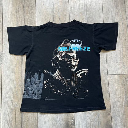 Batman &amp; Robin 1997 Mr. Freeze Promo Tee is a black T-shirt featuring a front graphic of Batman’s masked bust set against a grey city background, with a red Batman &amp; Robin logo above. The cityscape continues from the front around to the back, where a realistic Mr. Freeze graphic appears with his blue text incorporating the Batman symbol. Small 1997 licensing text is printed beneath the back graphic.