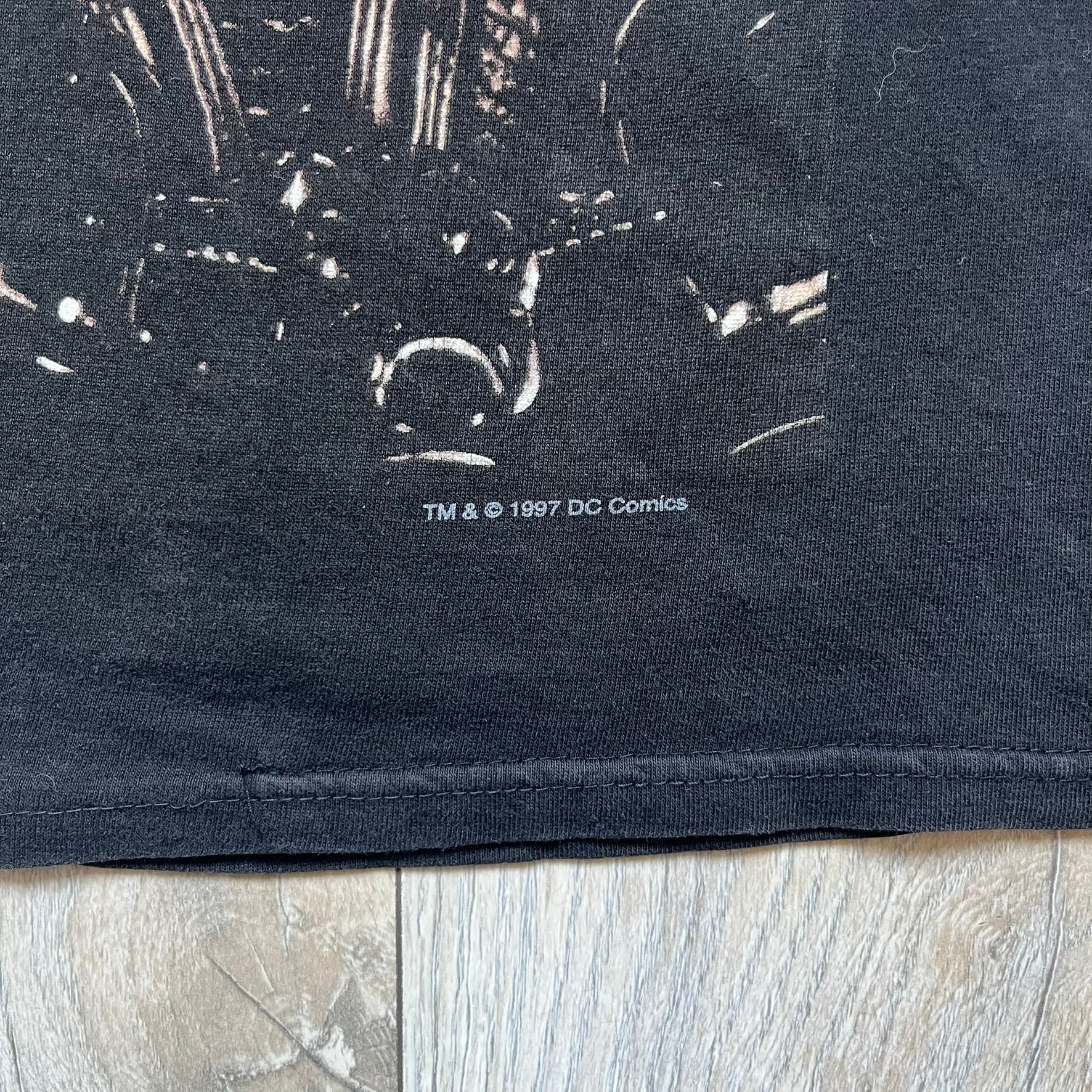 Batman &amp; Robin 1997 Mr. Freeze Promo Tee is a black T-shirt featuring a front graphic of Batman’s masked bust set against a grey city background, with a red Batman &amp; Robin logo above. The cityscape continues from the front around to the back, where a realistic Mr. Freeze graphic appears with his blue text incorporating the Batman symbol. Small 1997 licensing text is printed beneath the back graphic.