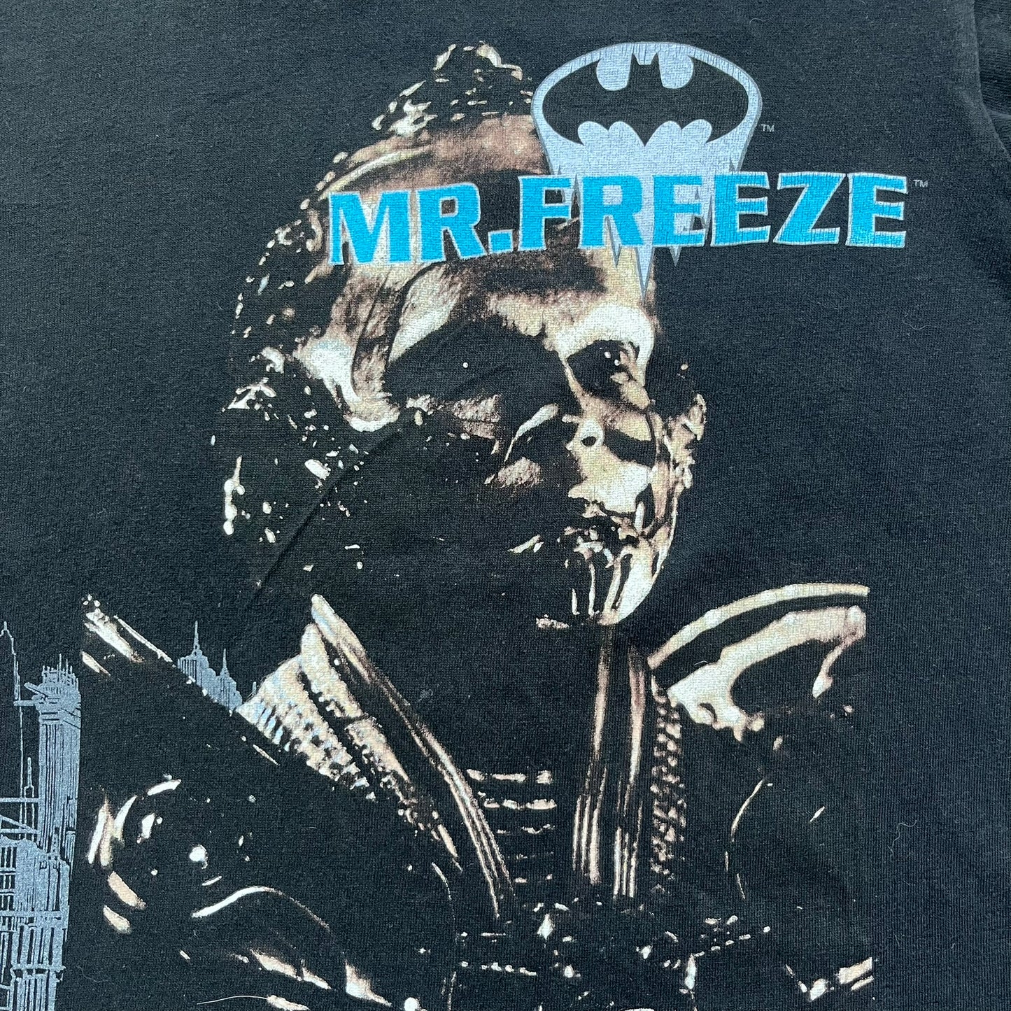 Batman &amp; Robin 1997 Mr. Freeze Promo Tee is a black T-shirt featuring a front graphic of Batman’s masked bust set against a grey city background, with a red Batman &amp; Robin logo above. The cityscape continues from the front around to the back, where a realistic Mr. Freeze graphic appears with his blue text incorporating the Batman symbol. Small 1997 licensing text is printed beneath the back graphic.