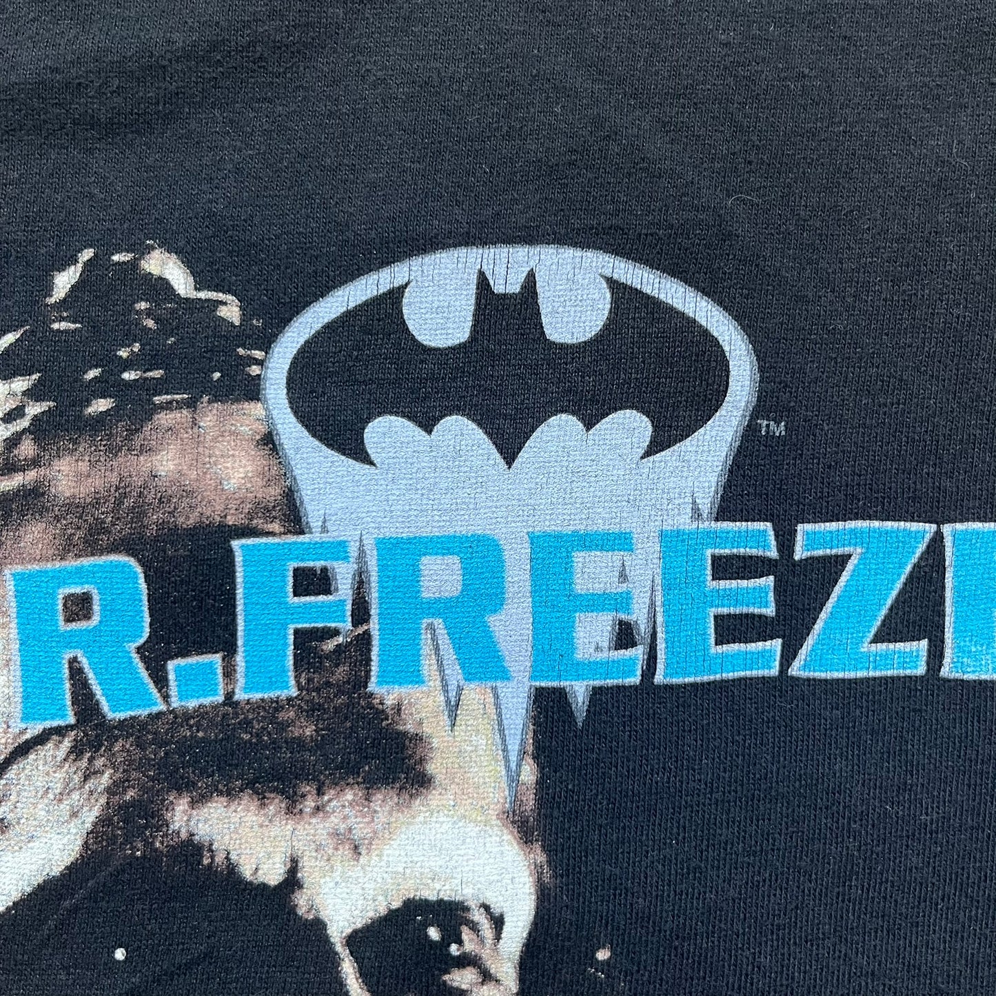 Batman &amp; Robin 1997 Mr. Freeze Promo Tee is a black T-shirt featuring a front graphic of Batman’s masked bust set against a grey city background, with a red Batman &amp; Robin logo above. The cityscape continues from the front around to the back, where a realistic Mr. Freeze graphic appears with his blue text incorporating the Batman symbol. Small 1997 licensing text is printed beneath the back graphic.