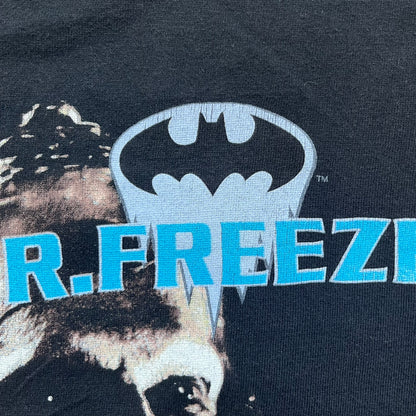 Batman &amp; Robin 1997 Mr. Freeze Promo Tee is a black T-shirt featuring a front graphic of Batman’s masked bust set against a grey city background, with a red Batman &amp; Robin logo above. The cityscape continues from the front around to the back, where a realistic Mr. Freeze graphic appears with his blue text incorporating the Batman symbol. Small 1997 licensing text is printed beneath the back graphic.