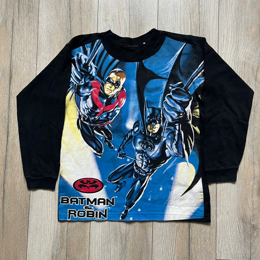 Batman &amp; Robin 1997 AOP Promo Longsleeve Tee is a black long sleeve T-shirt featuring a detailed all-over front print of Batman and Robin in a flying pose, created as part of the official promotional graphics for the film. The design includes a “Batman &amp; Robin” logo at the lower front with a small 1997 licensing print beneath it. The graphic is finished with a thick, paint-like application, giving it a textured, durable feel typical of vintage movie promo apparel.