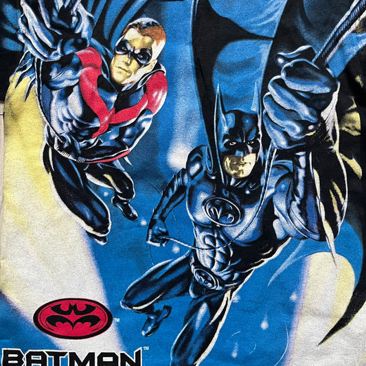 Batman &amp; Robin 1997 AOP Promo Longsleeve Tee is a black long sleeve T-shirt featuring a detailed all-over front print of Batman and Robin in a flying pose, created as part of the official promotional graphics for the film. The design includes a “Batman &amp; Robin” logo at the lower front with a small 1997 licensing print beneath it. The graphic is finished with a thick, paint-like application, giving it a textured, durable feel typical of vintage movie promo apparel.