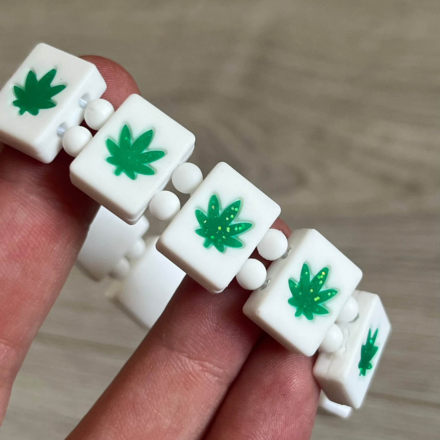 Bazo 1199 “420” Bracelet is a white silicone bead bracelet featuring glitter cannabis leaf graphics across the beads and a “420” emoji graphic on one bead. The interior includes a glitter red 1199 logo. It comes in the original plastic foil packaging.&nbsp;