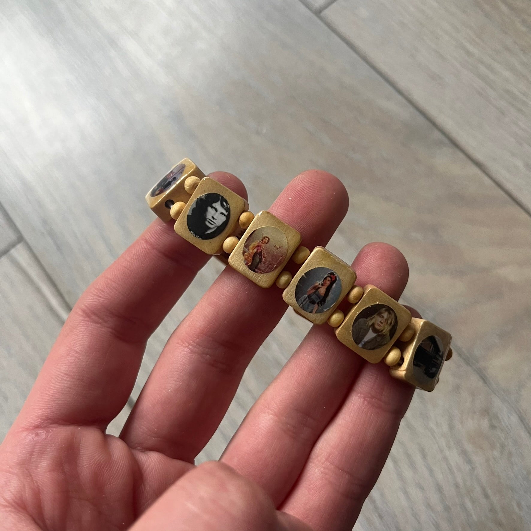 Beige “Forever 27 Club” bracelet by Bazo 1199, showcasing painted wooden beads and engraved portraits of legendary artists from the infamous 27 Club like Kurt Cobain, Basquiat, and Fredo Santana. Signed “forever 27 club” and “1199” on the inside.