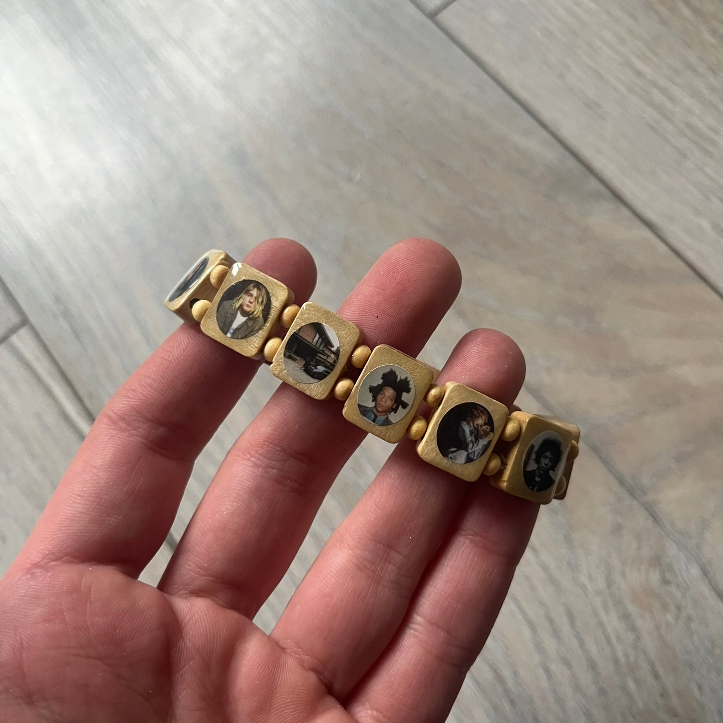 Beige “Forever 27 Club” bracelet by Bazo 1199, showcasing painted wooden beads and engraved portraits of legendary artists from the infamous 27 Club like Kurt Cobain, Basquiat, and Fredo Santana. Signed “forever 27 club” and “1199” on the inside.