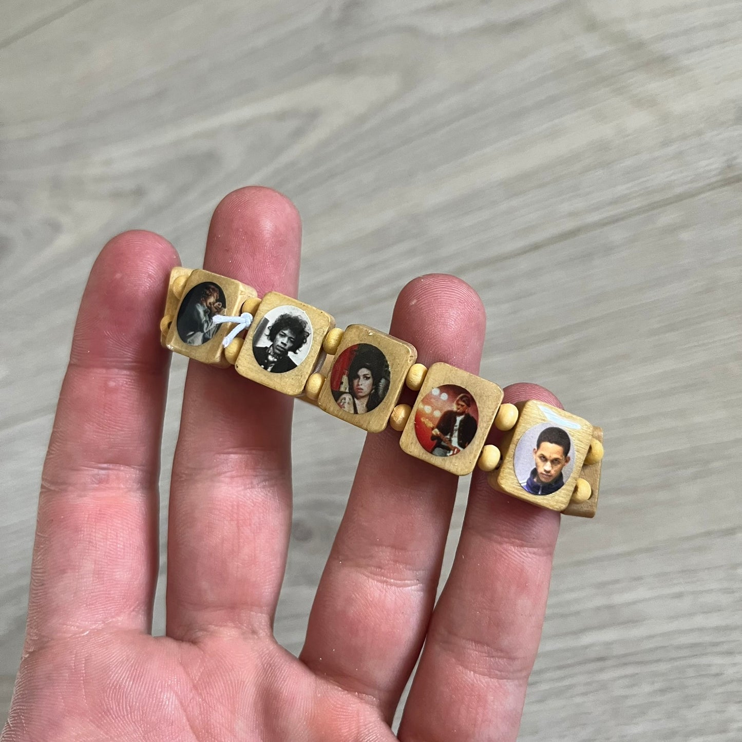 Beige “Forever 27 Club” bracelet by Bazo 1199, showcasing painted wooden beads and engraved portraits of legendary artists from the infamous 27 Club like Kurt Cobain, Basquiat, and Fredo Santana. Signed “forever 27 club” and “1199” on the inside.