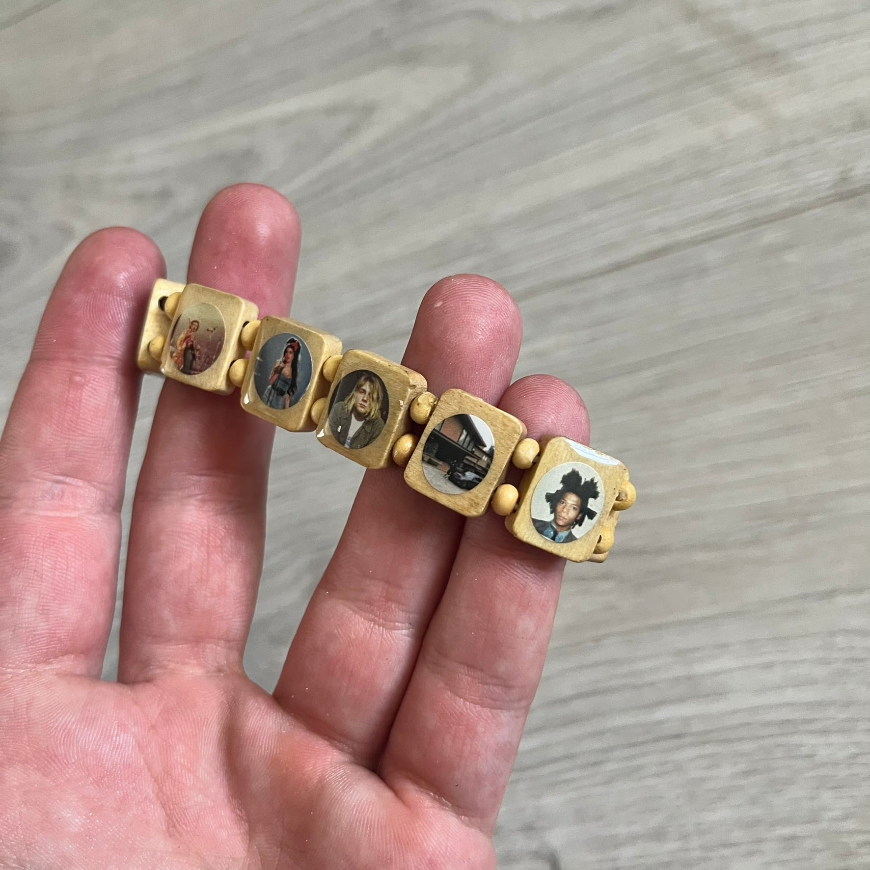 Beige “Forever 27 Club” bracelet by Bazo 1199, showcasing painted wooden beads and engraved portraits of legendary artists from the infamous 27 Club like Kurt Cobain, Basquiat, and Fredo Santana. Signed “forever 27 club” and “1199” on the inside.