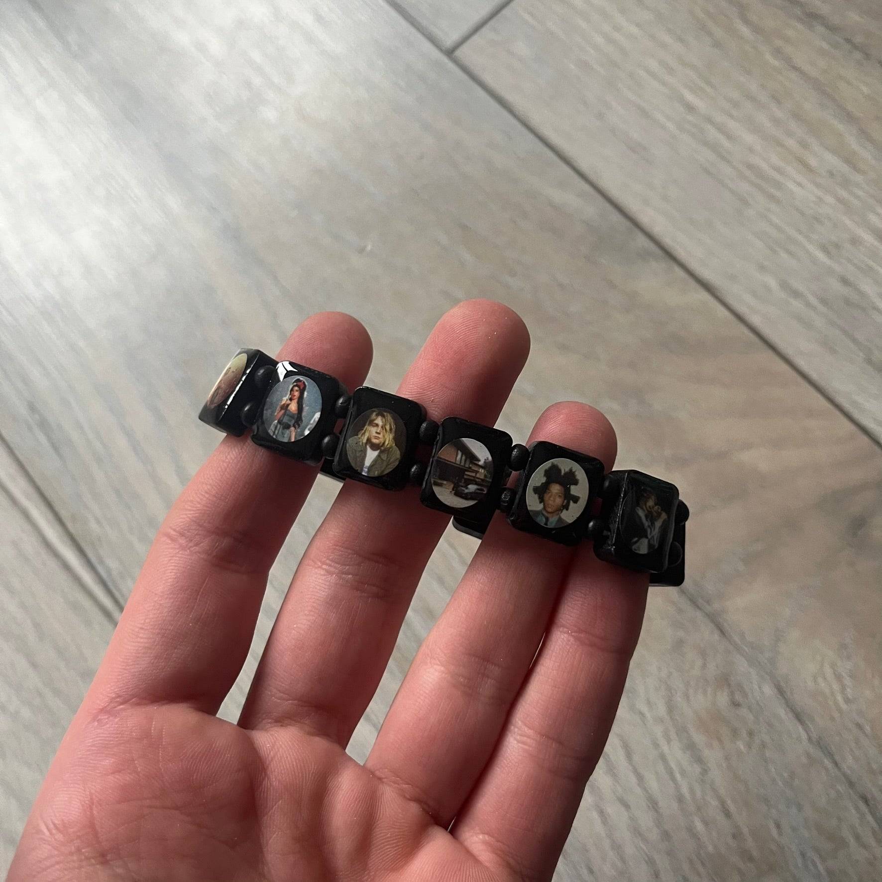 Black “Forever 27 Club” bracelet by Bazo 1199, showcasing painted wooden beads and engraved portraits of legendary artists from the infamous 27 Club like Kurt Cobain, Basquiat, and Fredo Santana. Signed “forever 27 club” and “1199” on the inside.