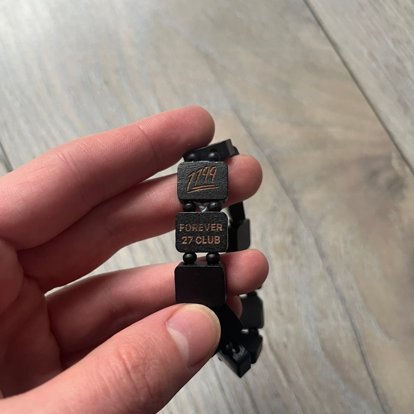 Black “Forever 27 Club” bracelet by Bazo 1199, showcasing painted wooden beads and engraved portraits of legendary artists from the infamous 27 Club like Kurt Cobain, Basquiat, and Fredo Santana. Signed “forever 27 club” and “1199” on the inside.