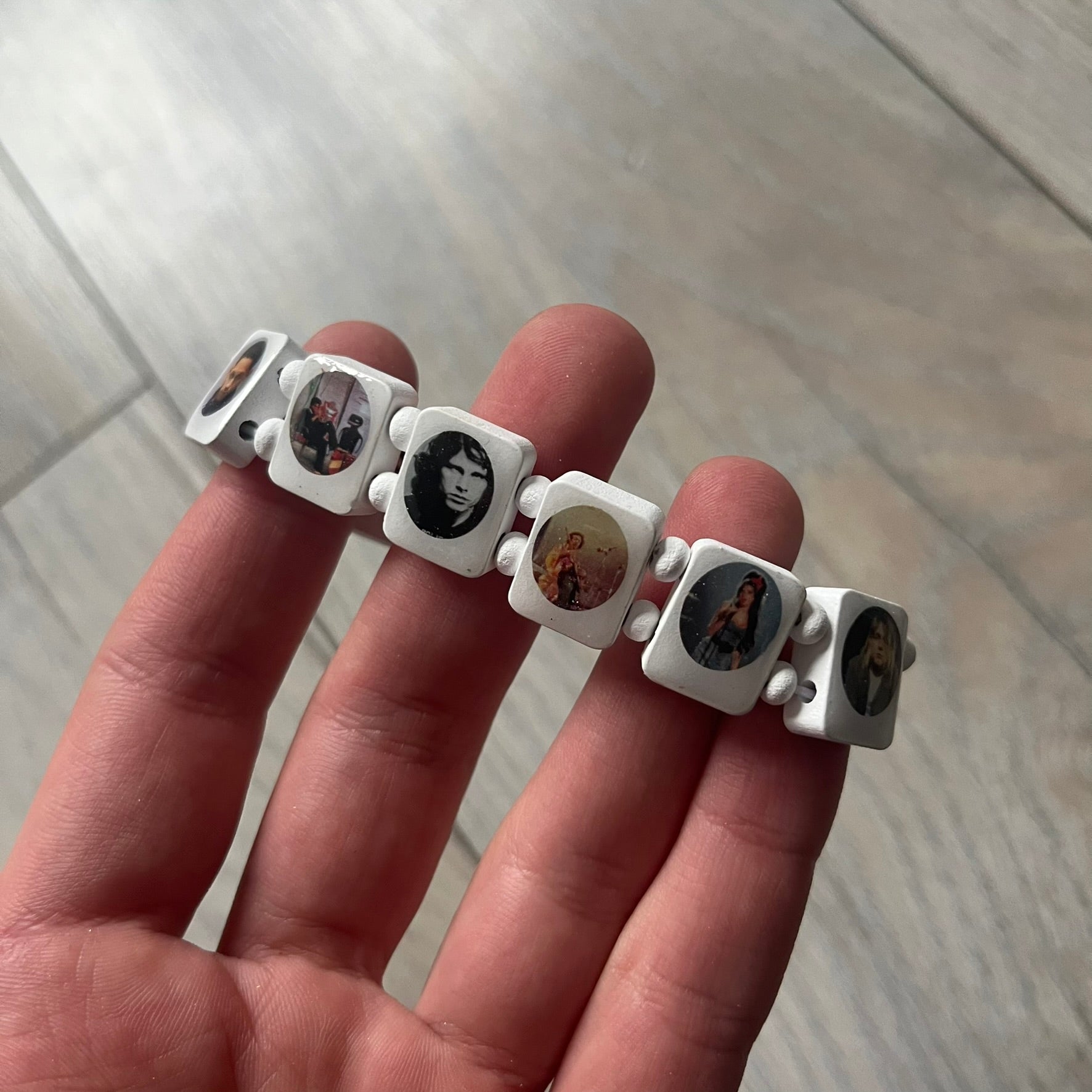 White “Forever 27 Club” bracelet by Bazo 1199, showcasing painted wooden beads and engraved portraits of legendary artists from the infamous 27 Club like Kurt Cobain, Basquiat, and Fredo Santana. Signed “forever 27 club” and “1199” on the inside.