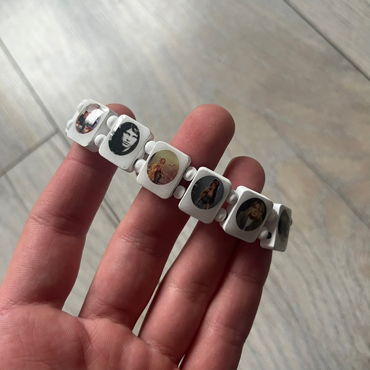 White “Forever 27 Club” bracelet by Bazo 1199, showcasing painted wooden beads and engraved portraits of legendary artists from the infamous 27 Club like Kurt Cobain, Basquiat, and Fredo Santana. Signed “forever 27 club” and “1199” on the inside.