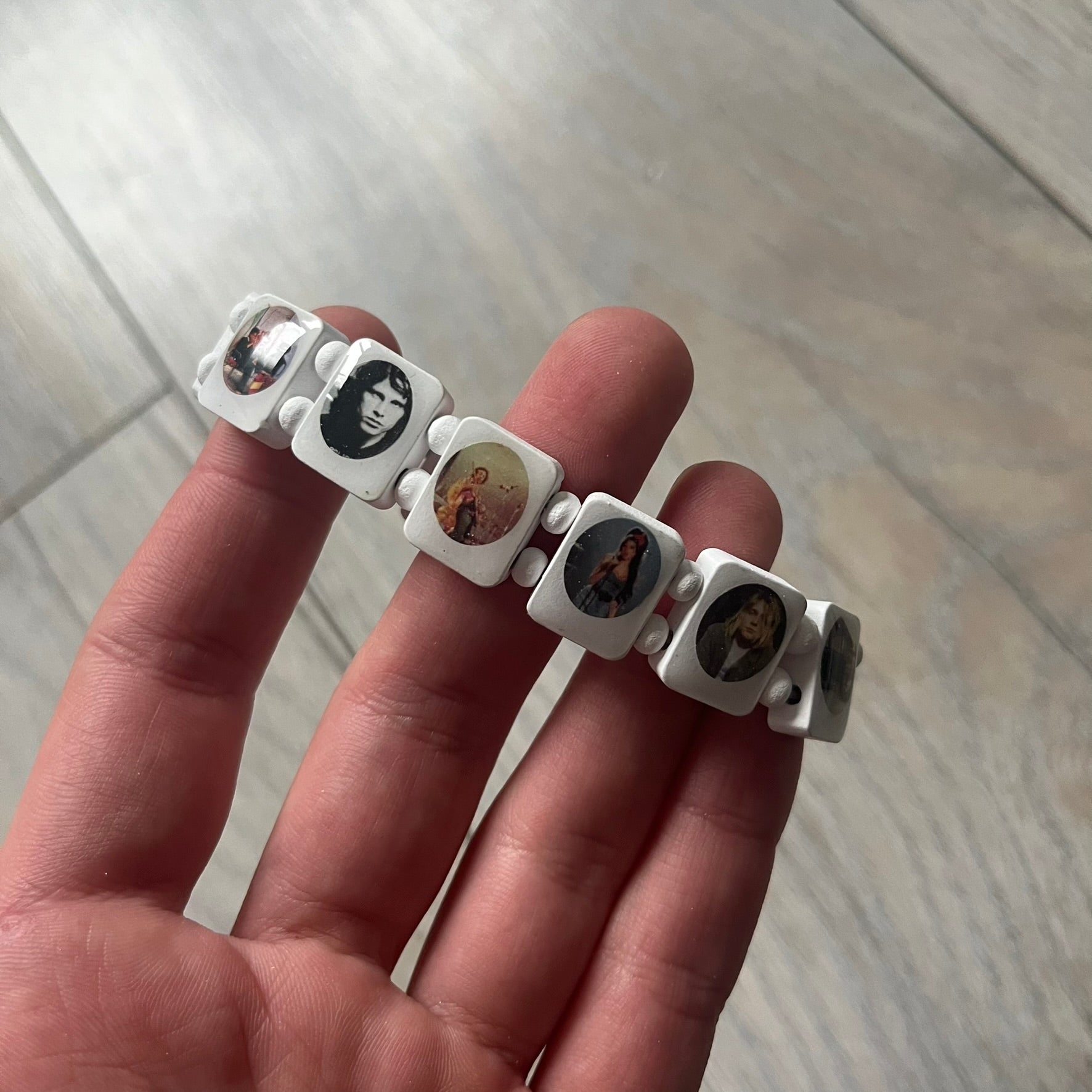 White “Forever 27 Club” bracelet by Bazo 1199, showcasing painted wooden beads and engraved portraits of legendary artists from the infamous 27 Club like Kurt Cobain, Basquiat, and Fredo Santana. Signed “forever 27 club” and “1199” on the inside.