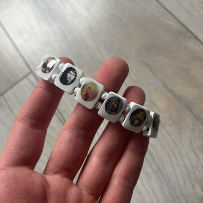 White “Forever 27 Club” bracelet by Bazo 1199, showcasing painted wooden beads and engraved portraits of legendary artists from the infamous 27 Club like Kurt Cobain, Basquiat, and Fredo Santana. Signed “forever 27 club” and “1199” on the inside.