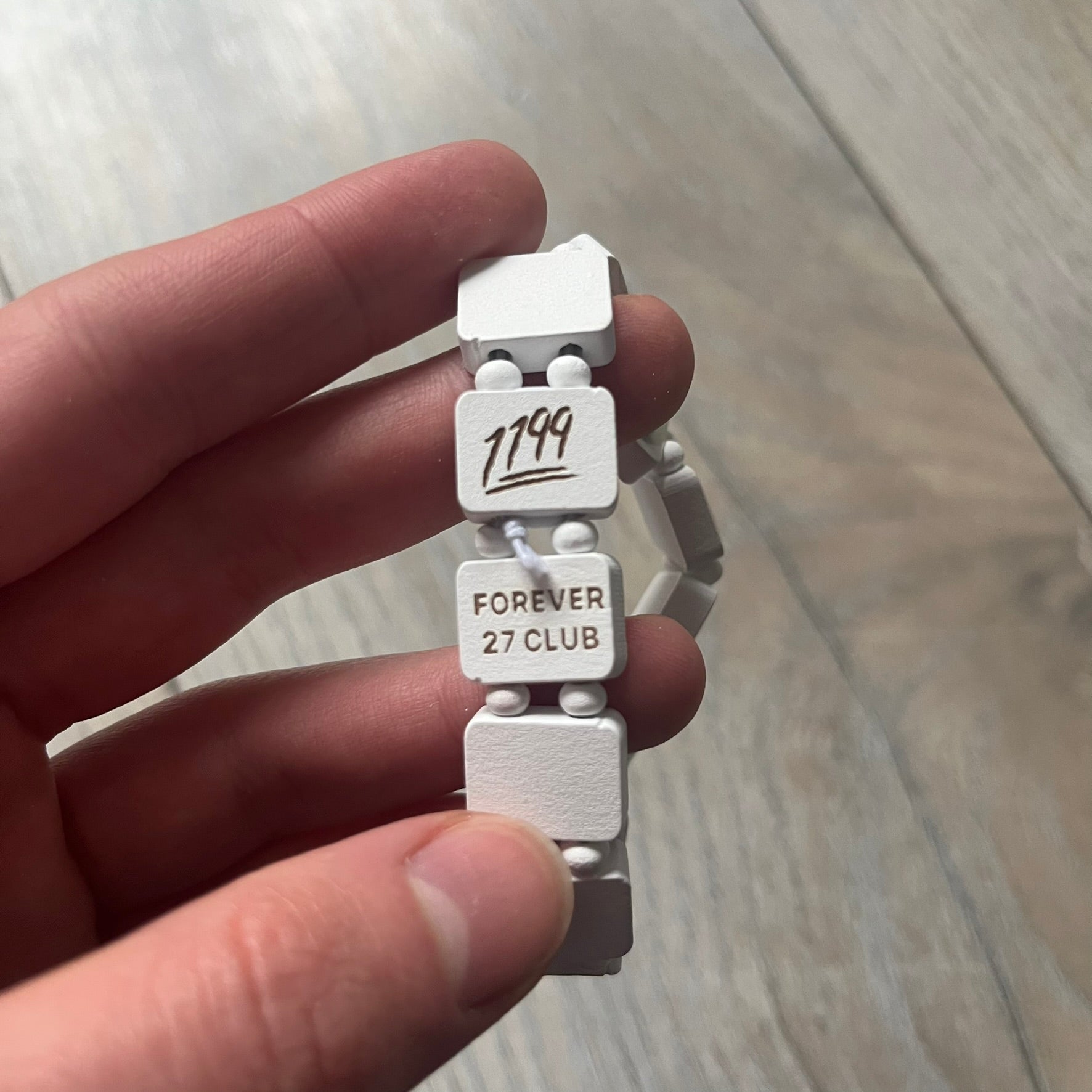 White “Forever 27 Club” bracelet by Bazo 1199, showcasing painted wooden beads and engraved portraits of legendary artists from the infamous 27 Club like Kurt Cobain, Basquiat, and Fredo Santana. Signed “forever 27 club” and “1199” on the inside.